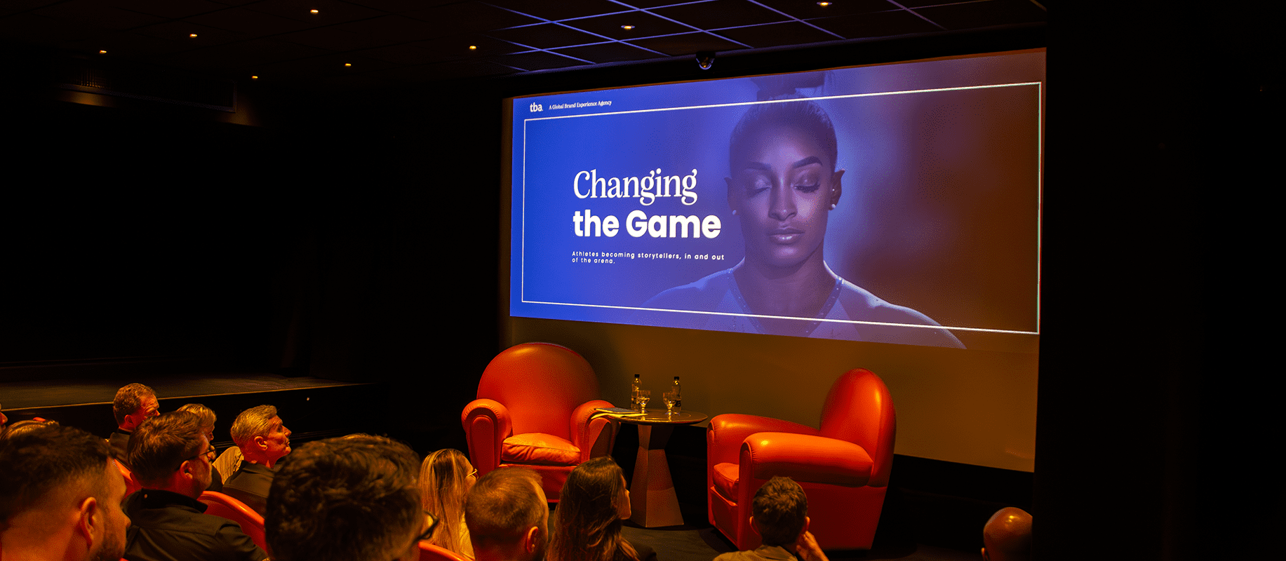 Audience in a small theatre watches a presentation slide titled ‘Changing the Game’ with a woman’s portrait; two red armchairs and a small table on stage.